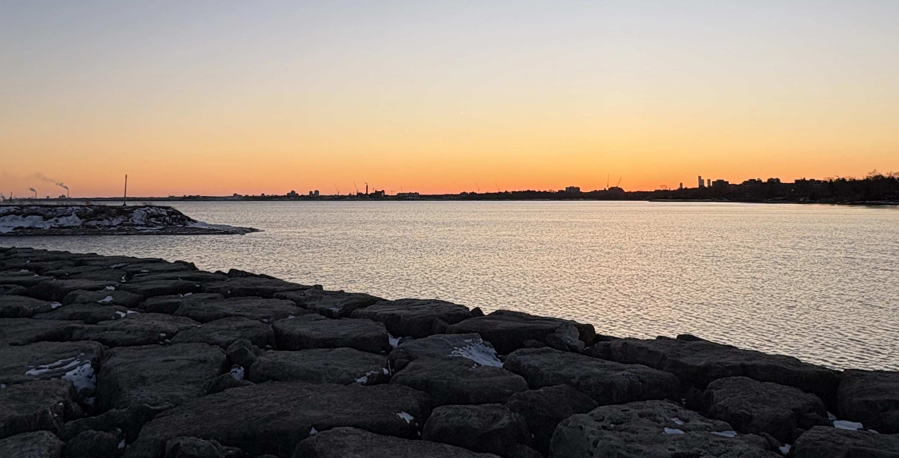 Sunset over Lake Ontario from the rocky shoreline at Colonel Samuel Smith Park in winter, with the distant Toronto skyline on the horizon